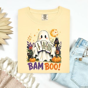 May include: A pale yellow t-shirt featuring a graphic of a white ghost holding three bamboo tiles. The design includes a black cat, orange pumpkins, a dark house, and a full moon. The text BAM BOO! is printed in purple and orange below the ghost.