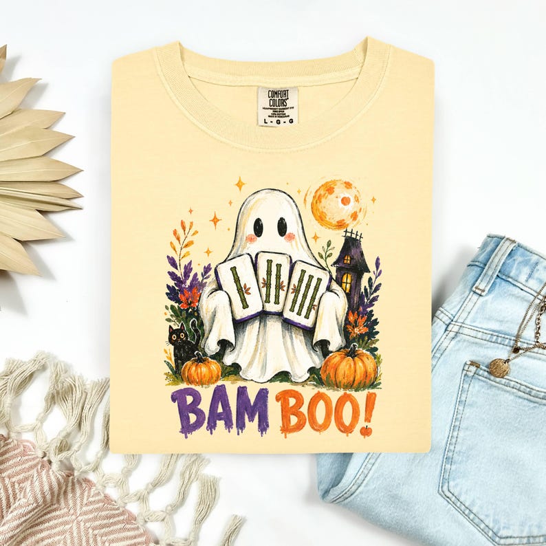 May include: A pale yellow t-shirt featuring a graphic of a white ghost holding three bamboo tiles. The design includes a black cat, orange pumpkins, a dark house, and a full moon. The text BAM BOO! is printed in purple and orange below the ghost.