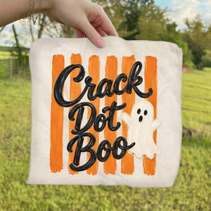 May include: A cream-colored fabric square featuring vertical orange painted stripes. The black text reads Crack Dot Boo in a cursive script. A small, white, hand-painted ghost with two black eyes and an open mouth is positioned to the right of the text.