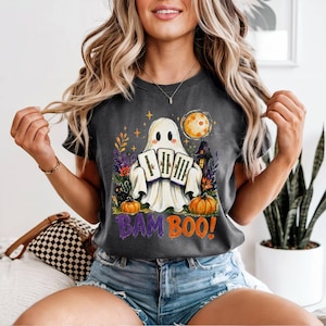 May include: A charcoal gray t-shirt featuring a graphic of a white ghost holding three bamboo stalks. The design includes a yellow moon, orange pumpkins, a black cat, and colorful foliage. The text BAM BOO is printed in purple and orange.