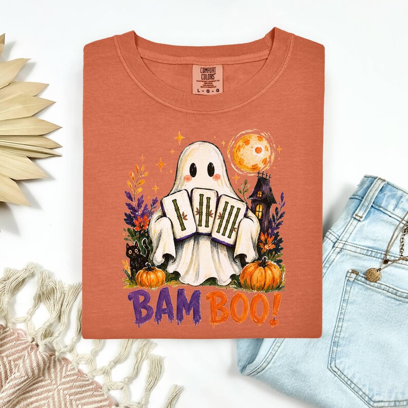 May include: A burnt orange t-shirt featuring a graphic of a white ghost holding three bamboo mahjong tiles. The design includes a black cat, pumpkins, a full moon, and a haunted house. The text BAM BOO is printed in purple and orange below.