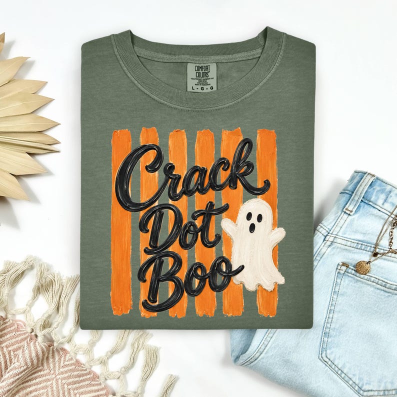 May include: An olive green t-shirt featuring a graphic of vertical orange brushstrokes behind the black text "Crack Dot Boo" and a small white ghost illustration. The shirt has a crew neckline and a Comfort Colors brand label at the inner neck.
