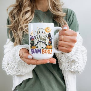 May include: A white ceramic mug featuring a graphic of a ghost holding three tarot cards, surrounded by pumpkins, flowers, and a haunted house. The text BAM BOO! is printed in purple and orange letters at the bottom of the design on the mug.