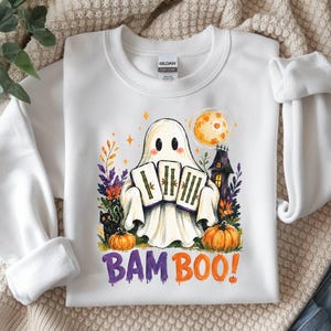 May include: A white crewneck sweatshirt featuring a graphic of a ghost holding three mahjong tiles. The design includes a black cat, pumpkins, a haunted house, and a full moon. The text BAM BOO! is printed in purple and orange below the illustration.