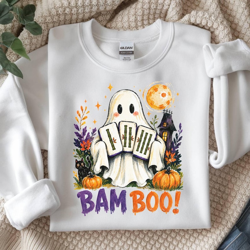 May include: A white crewneck sweatshirt featuring a graphic of a ghost holding three mahjong tiles. The design includes a black cat, pumpkins, a haunted house, and a full moon. The text BAM BOO! is printed in purple and orange below the illustration.