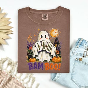 May include: A brown t-shirt featuring a graphic of a white ghost holding three bamboo tiles. The design includes a black cat, orange pumpkins, a full moon, and a haunted house. The text BAM BOO is printed in purple and orange below the ghost.