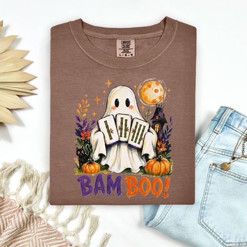 May include: A brown t-shirt featuring a graphic of a white ghost holding three bamboo tiles. The design includes a black cat, orange pumpkins, a full moon, and a haunted house. The text BAM BOO is printed in purple and orange below the ghost.