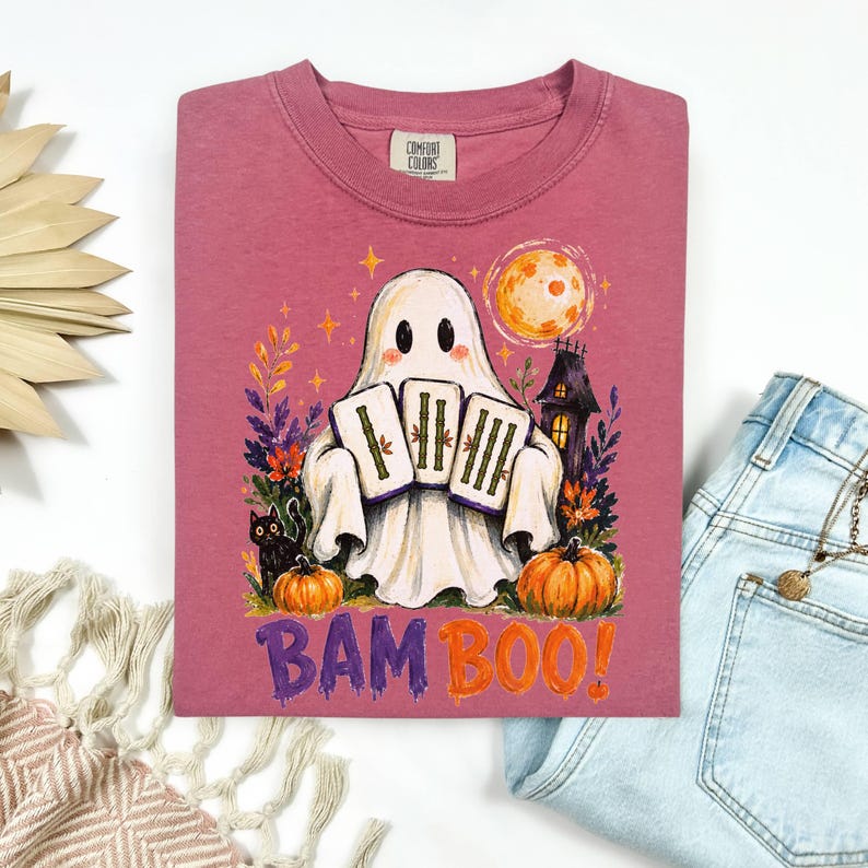 May include: A dusty rose t-shirt featuring a graphic of a white ghost holding three mahjong tiles. The design includes a black cat, orange pumpkins, a yellow moon, and a haunted house. The text BAMBOO is printed in purple and orange below the ghost.