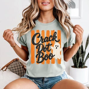 May include: A light green t-shirt featuring a graphic with vertical orange brushstroke stripes and the black text "Crack Dot Boo" written in a cursive font. A small, white, cartoon-style ghost with a surprised expression is positioned to the right.