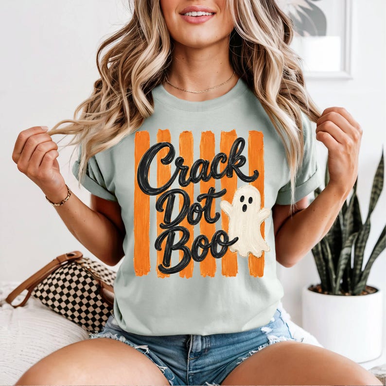 May include: A light green t-shirt featuring a graphic with vertical orange brushstroke stripes and the black text "Crack Dot Boo" written in a cursive font. A small, white, cartoon-style ghost with a surprised expression is positioned to the right.