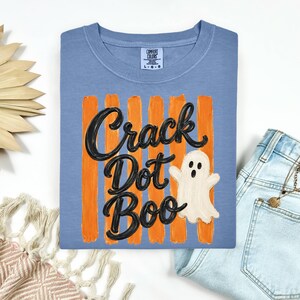 May include: A blue short-sleeved t-shirt featuring a graphic of vertical orange brushstrokes, the black text "Crack Dot Boo", and a white ghost illustration. The shirt has a Comfort Colors brand label on the inside of the crew neckline.