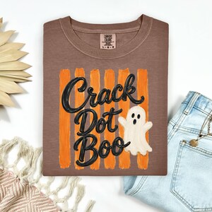 May include: A brown t-shirt featuring a graphic with vertical orange brushstrokes and the black text Crack Dot Boo. A small white ghost illustration is positioned to the right of the text. The shirt has a Comfort Colors brand label at the neck.