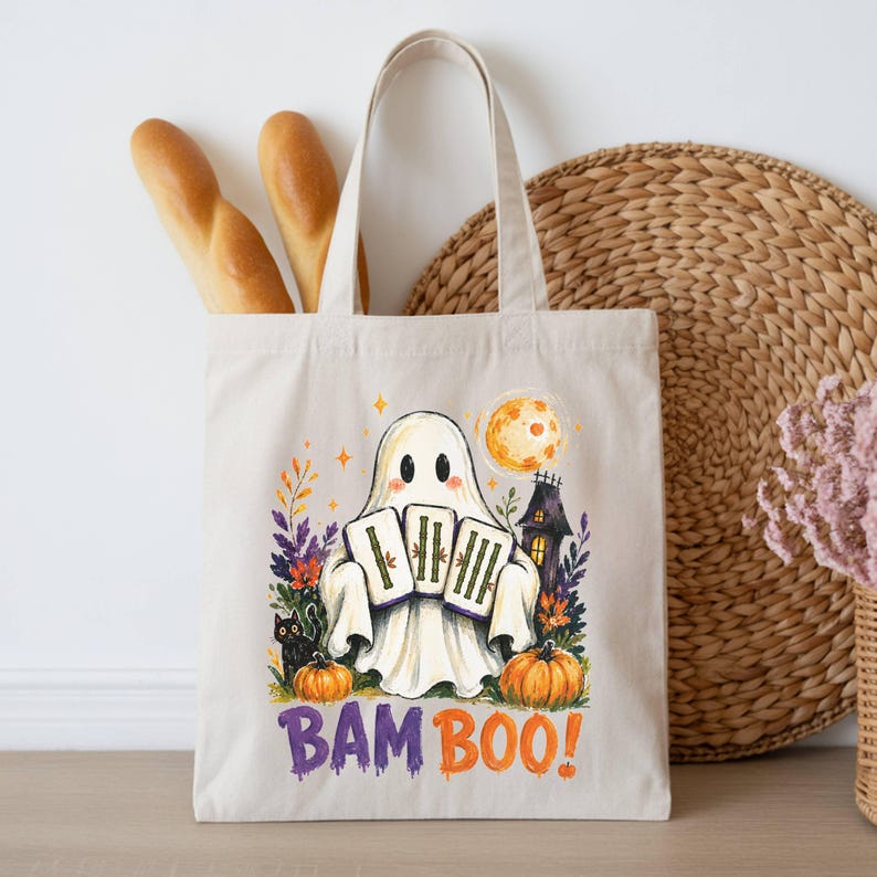 May include: A beige canvas tote bag featuring a colorful graphic of a ghost holding three bamboo tiles. The design includes a black cat, pumpkins, a full moon, and a haunted house. The text BAM BOO! is printed in purple and orange at the bottom.