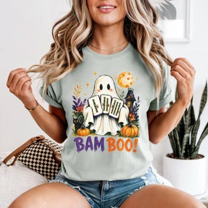 May include: A light green t-shirt featuring a graphic of a white ghost holding three bamboo stalks. The design includes a black cat, orange pumpkins, a yellow moon, and a dark house. The text BAM BOO! is printed in purple and orange below the ghost.