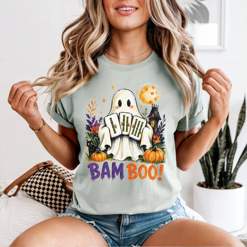 May include: A light green t-shirt featuring a graphic of a white ghost holding three bamboo stalks. The design includes a black cat, orange pumpkins, a yellow moon, and a dark house. The text BAM BOO! is printed in purple and orange below the ghost.