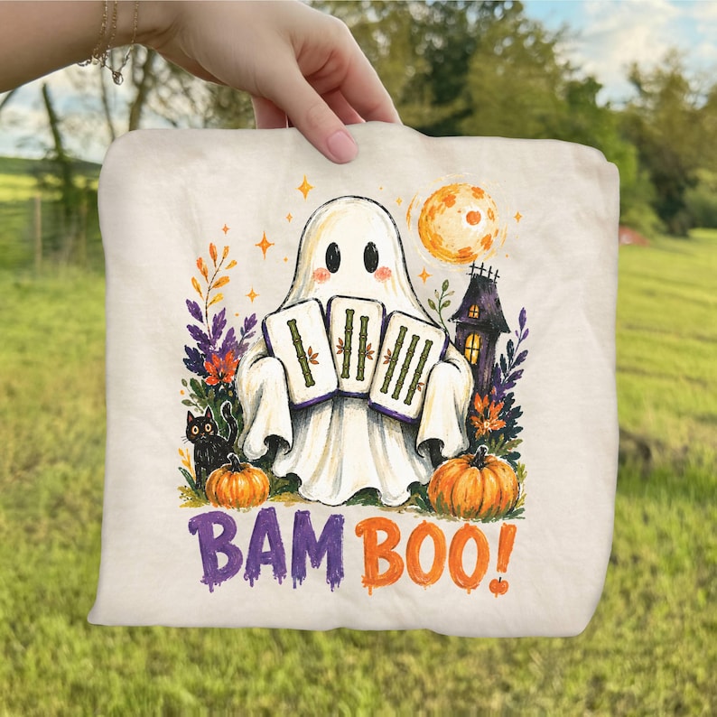 May include: A cream-colored fabric item featuring a graphic of a white ghost holding three bamboo tiles. The design includes a black cat, orange pumpkins, a dark house, and a full moon. The text BAM BOO! is printed in purple and orange below.