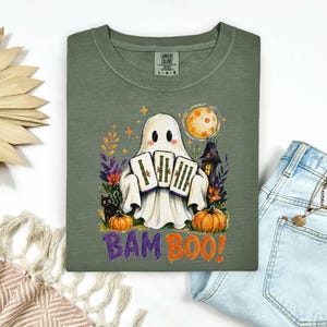 May include: An olive green t-shirt featuring a graphic of a ghost holding three bamboo stalks. The design includes a black cat, two orange pumpkins, a full moon, a dark house, and colorful foliage. The text BAM BOO is printed in purple and orange.