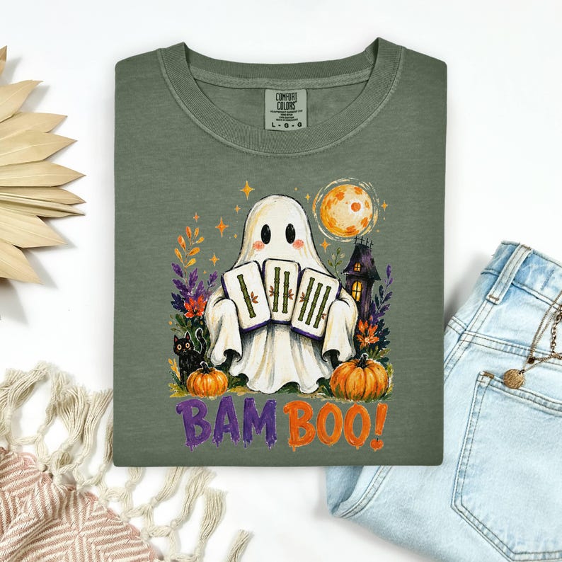 May include: An olive green t-shirt featuring a graphic of a ghost holding three bamboo stalks. The design includes a black cat, two orange pumpkins, a full moon, a dark house, and colorful foliage. The text BAM BOO is printed in purple and orange.