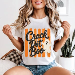 May include: A white short-sleeved t-shirt featuring a graphic print of vertical orange brushstrokes. Overlaid on the stripes is the black text "Crack Dot Boo" in a cursive font, accompanied by a small, off-white cartoon ghost illustration.