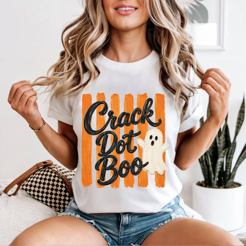 May include: A white short-sleeved t-shirt featuring a graphic print of vertical orange brushstrokes. Overlaid on the stripes is the black text "Crack Dot Boo" in a cursive font, accompanied by a small, off-white cartoon ghost illustration.