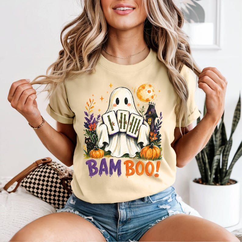 May include: A light yellow t-shirt featuring a graphic of a white ghost holding three bamboo stalks. The design includes a black cat, two orange pumpkins, a dark house, and a full moon. The text BAM BOO! is printed in purple and orange letters.
