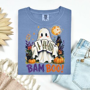 May include: A blue t-shirt featuring a graphic of a white ghost holding three mahjong tiles. The design includes a black cat, orange pumpkins, a full moon, and a haunted house. The text BAM BOO is printed in purple and orange below the ghost.