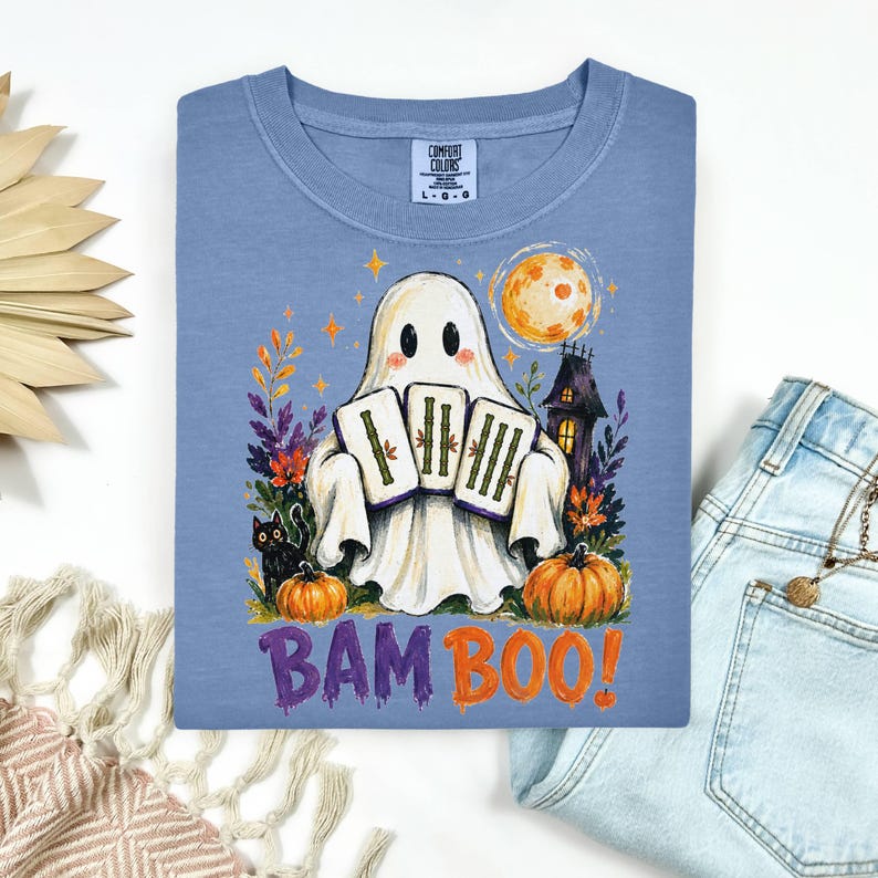May include: A blue t-shirt featuring a graphic of a white ghost holding three mahjong tiles. The design includes a black cat, orange pumpkins, a full moon, and a haunted house. The text BAM BOO is printed in purple and orange below the ghost.