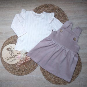 Könnte beinhalten: Ein Baby-Outfit-Set mit einem weißen Langarmshirt mit Rüschenärmeln und einem hellvioletten Latzkleid. Das Shirt hat eine gerippte Textur. Das Kleid hat Holzknöpfe an den Trägern. Das Set wird auf einer gewebten Matte präsentiert.