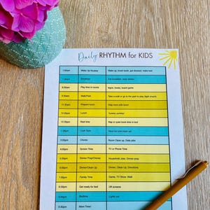 May include: A printed daily routine chart for kids, featuring a schedule from 7:00 am to 9:30 pm. The chart has a yellow and blue color scheme with the text "Daily RHYTHM for KIDS". A pen rests on the chart, and a vase of pink flowers is in the background.