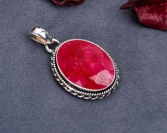 May include: A silver pendant featuring a large, oval-shaped, vivid red gemstone. The gemstone is framed by a twisted rope design. The pendant has a silver loop. The background is a dark grey.