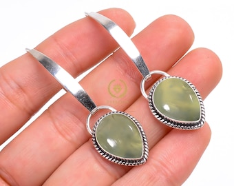 May include: A pair of silver earrings with a teardrop-shaped, pale green stone pendant. The stone is framed by a silver rope-like border. The earrings have a long, flat silver hook.