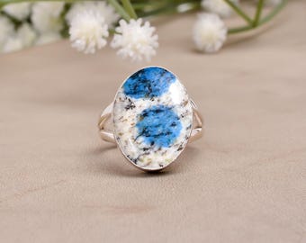 May include: Oval-shaped ring featuring a k2 stone with blue and white patterns. The ring has a silver band with a split shank design. The gemstone is the focal point, set against a neutral background with floral accents.