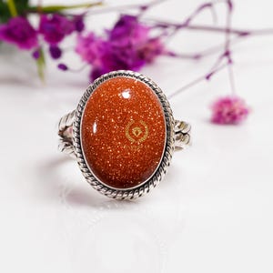 May include: A silver ring featuring a large oval-shaped red sunstone. The stone is set in a silver bezel with a rope-like design. The stone has a sparkly, glittery texture.