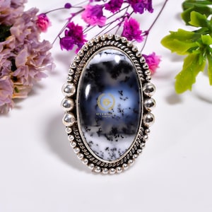 Pode incluir: Um anel de prata com uma grande pedra oval de ágata dendrítica. A pedra é uma mistura de branco, preto e cinza com um padrão de redemoinho. O anel tem uma borda com contas.