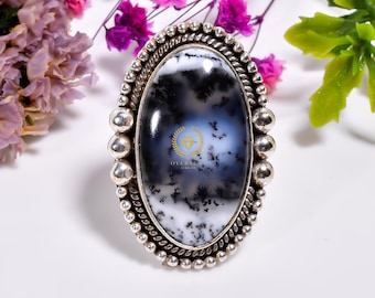 Puede incluir: Un anillo de plata con una gran piedra ovalada de ágata dendrítica. La piedra es una mezcla de blanco, negro y gris con un patrón de remolino. El anillo tiene un borde con cuentas.