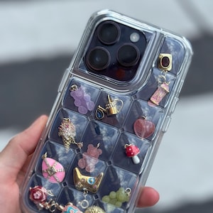 May include: Clear phone case with a grid of compartments, each holding a charm. Charms include a purple gummy bear, a gold watering can, a pink egg, a red mushroom, and a gold crown. The case is held by a hand.