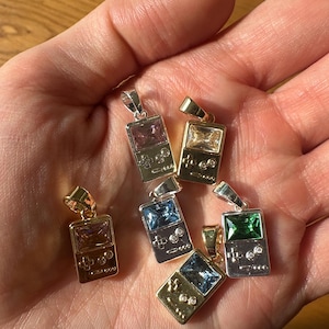 May include: A collection of silver and gold-toned pendant necklaces. Each pendant is shaped like a retro handheld game console, featuring colorful rectangular gemstones in pink, blue, green, and clear. The pendants are displayed on a light brown surface.