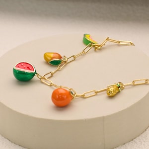 Könnte beinhalten: Eine goldene Kettenkette mit Anhängern in Form von Wassermelone, Orange, Ananas, Mango und einer Scheibe Wassermelone. Die Anhänger sind mit Emaille in leuchtenden Farben bemalt.