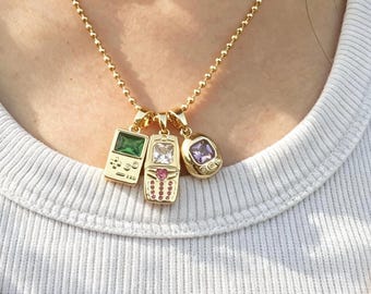 May include: A gold-toned ball chain necklace featuring three distinct charms: a rectangular handheld game console pendant with a green stone, a vintage mobile phone pendant with a clear and pink stone, and an oval digital pet pendant with a purple stone.