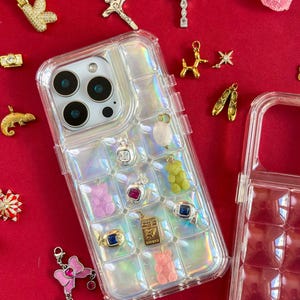 May include: A clear iridescent phone case with various charms, including a pink bow, a gold dog, and a gummy bear. Other charms include a cross, a ring with a blue stone, and a gold pendant with the word "LOWES".
