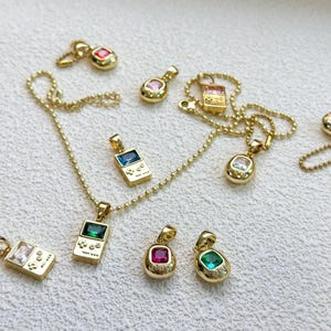 May include: A collection of gold-coloured pendant necklaces and charms with colourful gemstone details. The pendants include a handheld game console design and oval shapes. The gemstones are in shades of red, blue, green, and pink.