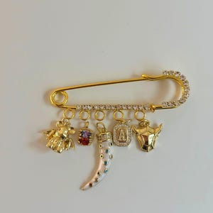 May include: A gold-colored safety pin with a row of clear rhinestones along the curved end. The pin features several dangling charms, including a bear, a gem, a horn, a pendant with the letter "A", and a bull's head.