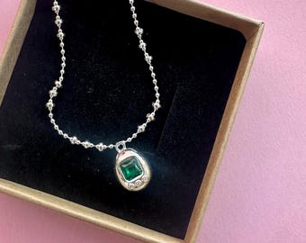 May include: A silver necklace with a pendant featuring a square, emerald-green gemstone. The pendant is surrounded by a silver frame and small, clear stones. The necklace is displayed in a brown box with a black interior, set against a pink background.