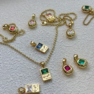 Puede incluir: Collares con colgantes dorados con detalles de gemas de colores. La colección incluye colgantes con forma de consolas de juegos portátiles y gemas ovaladas en varios colores, como rosa, rojo, verde y azul. Se exhiben sobre una superficie neutra.