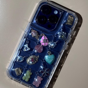 May include: A clear phone case with a blue interior, filled with various charms. Charms include a pink teardrop, a green heart, a flower, and two gummy bear charms. The case is designed to hold multiple charms.