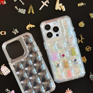 Puede incluir: Dos fundas transparentes para teléfono con un patrón de cuadrícula, una llena de amuletos de colores. Los amuletos incluyen un perro dorado, un osito de goma, una cruz y una cámara. Varios otros amuletos están esparcidos sobre una superficie negra.
