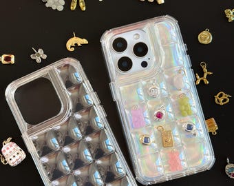 May include: Two clear phone cases with a grid pattern, one filled with colorful charms. Charms include a gold dog, a gummy bear, a cross, and a camera. Various other charms are scattered around the cases on a black surface.