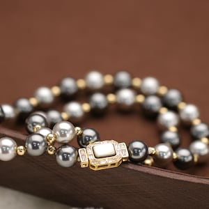 以下が含まれることがあります： 黒と白の真珠でできた、2連の真珠のネックレス。金色の留め具には、小さな石で囲まれた正方形の石が飾られています。