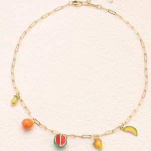 Könnte beinhalten: Eine goldene Kettenkette mit bunten Fruchtanhängern. Die Anhänger umfassen eine Ananas, eine Orange, eine Wassermelone, eine Mango und eine regenbogenfarbene Halbmond.