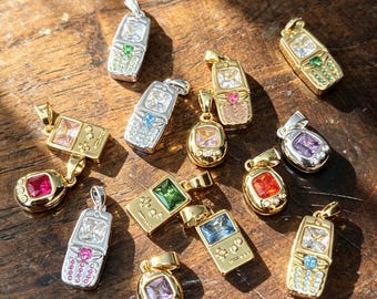 May include: A collection of metal pendants shaped like retro handheld gaming consoles and digital pets, featuring various metallic finishes including gold and silver tones, accented with colorful faceted gemstones set into the screens and buttons.